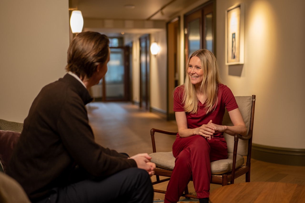 Behandler i burgunder farget uniform sitter i samtale med pasient i lounget hos Vinci Dental Studio